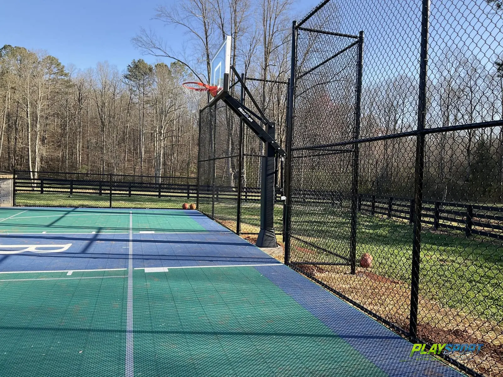 Corner of a green and blue modular backyard basketball court with a black chain-link fence enclosure