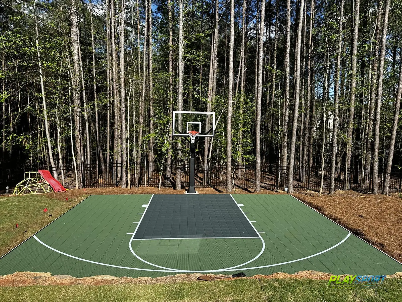 Compact green modular backyard basketball court against a pine forest