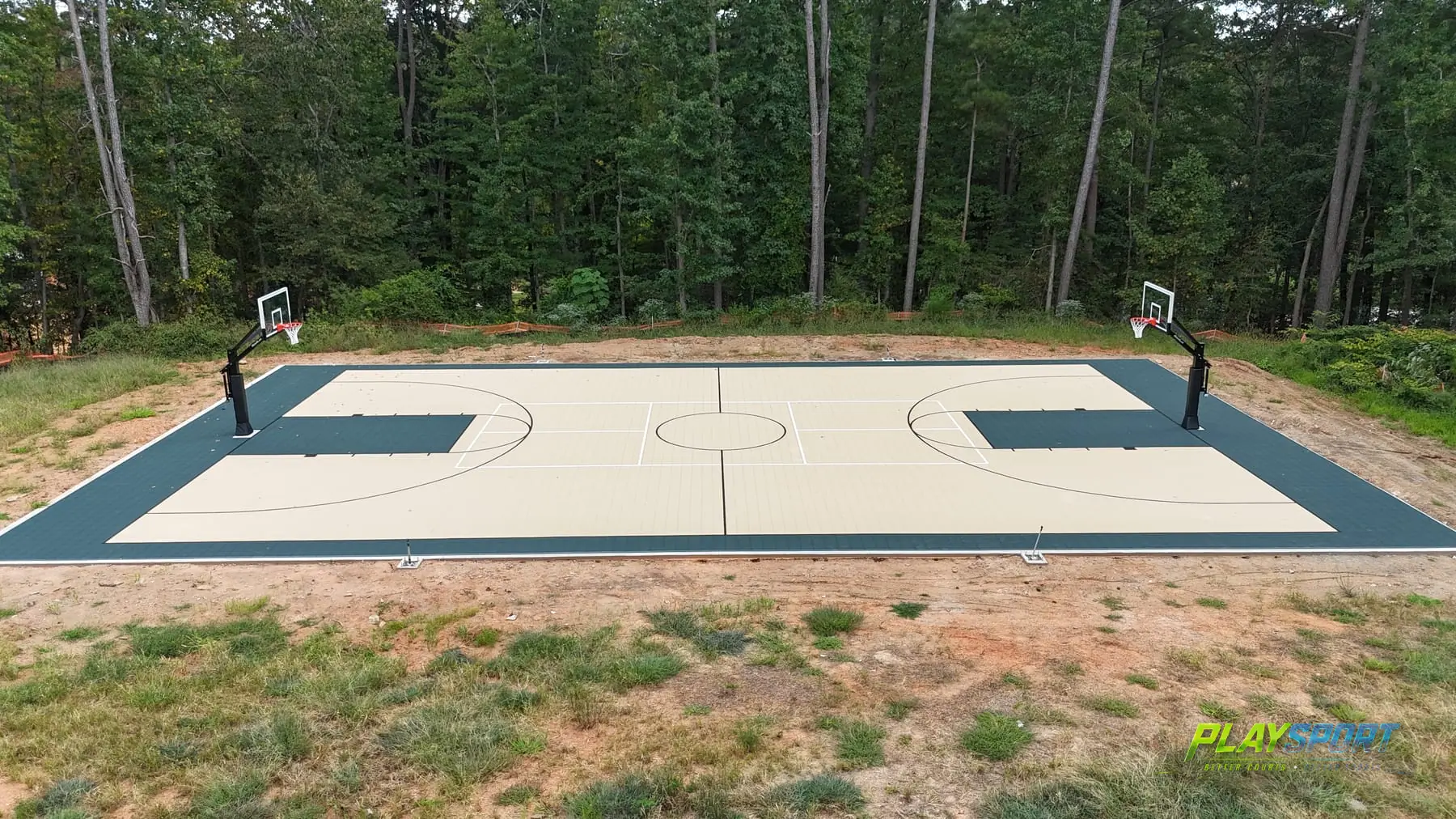 Full-size basketball court with a sand-tan surface and forest green trim, two hoops, surrounded by pine woods