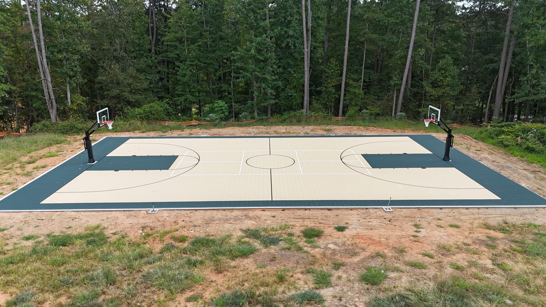 Full-size basketball court with sand-tan surface and forest green trim — two hoops, surrounded by pine woods