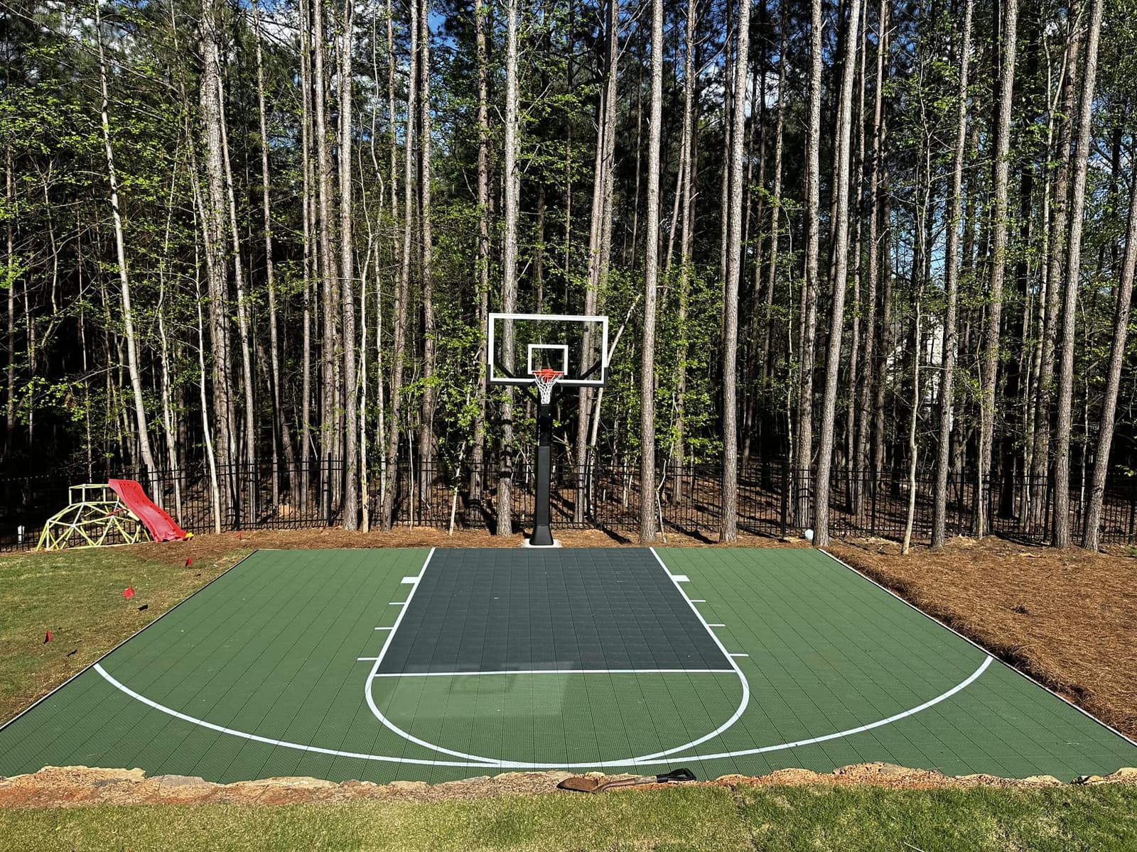 Compact green backyard basketball court against a pine forest