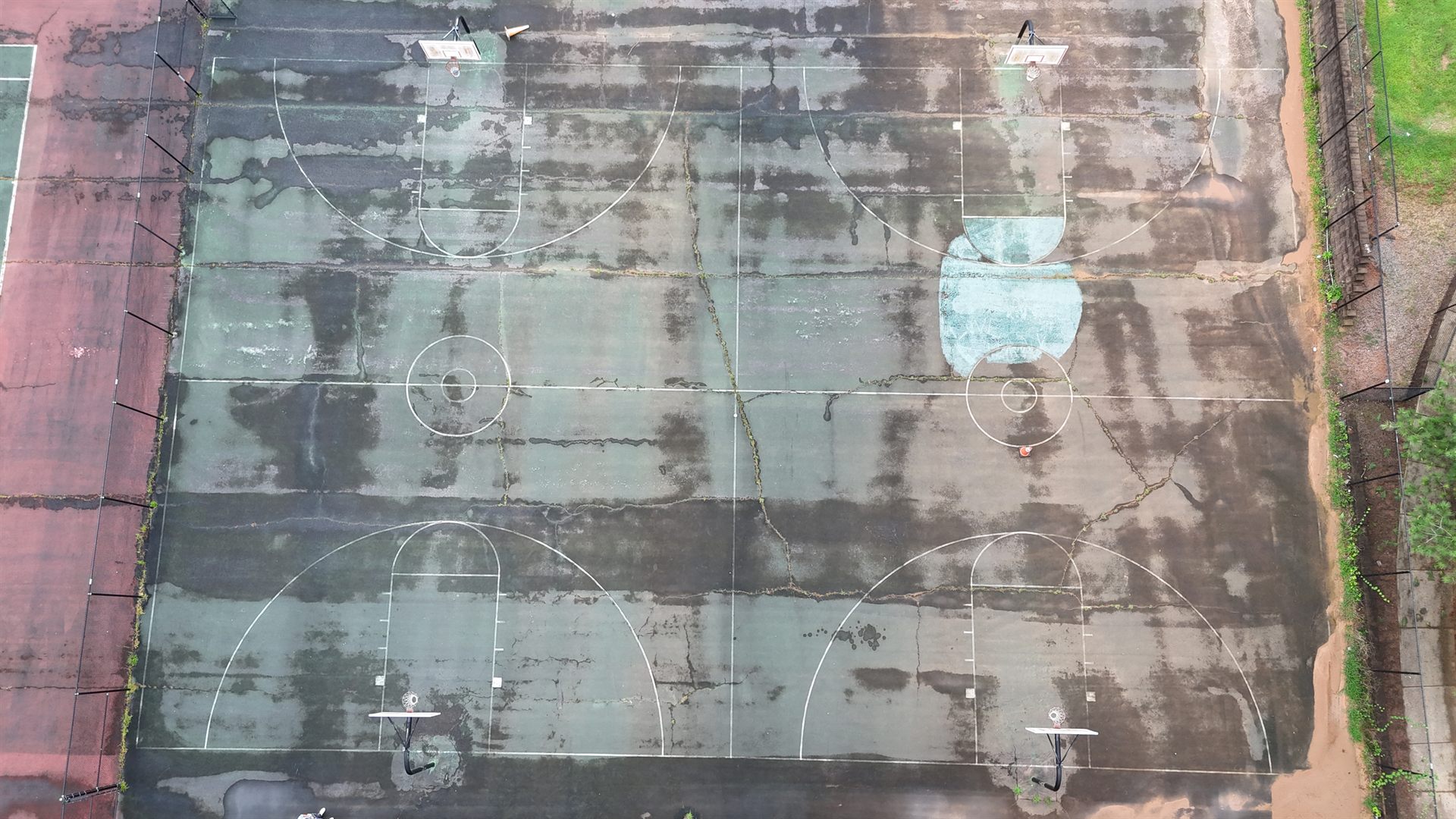 Cracked, faded painted-acrylic basketball court with weeds growing through the seams — the kind of surface PlaySport modular tiles replace