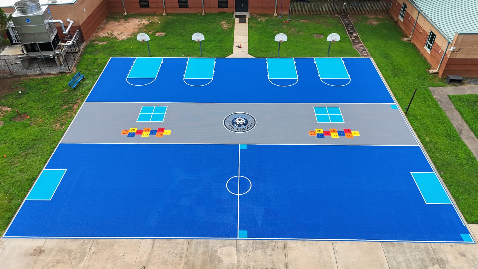 Aerial view of the finished Summit Hill Elementary playground — blue PlaySport multi-sport tile courts with basketball, pickleball, four square, hopscotch and a soccer field around a custom Husky center logo
